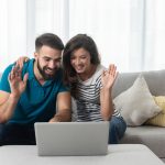 Young,Couple,Sitting,In,Their,Living,Room,With,Laptop,Waving
