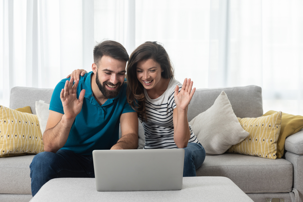 Young,Couple,Sitting,In,Their,Living,Room,With,Laptop,Waving